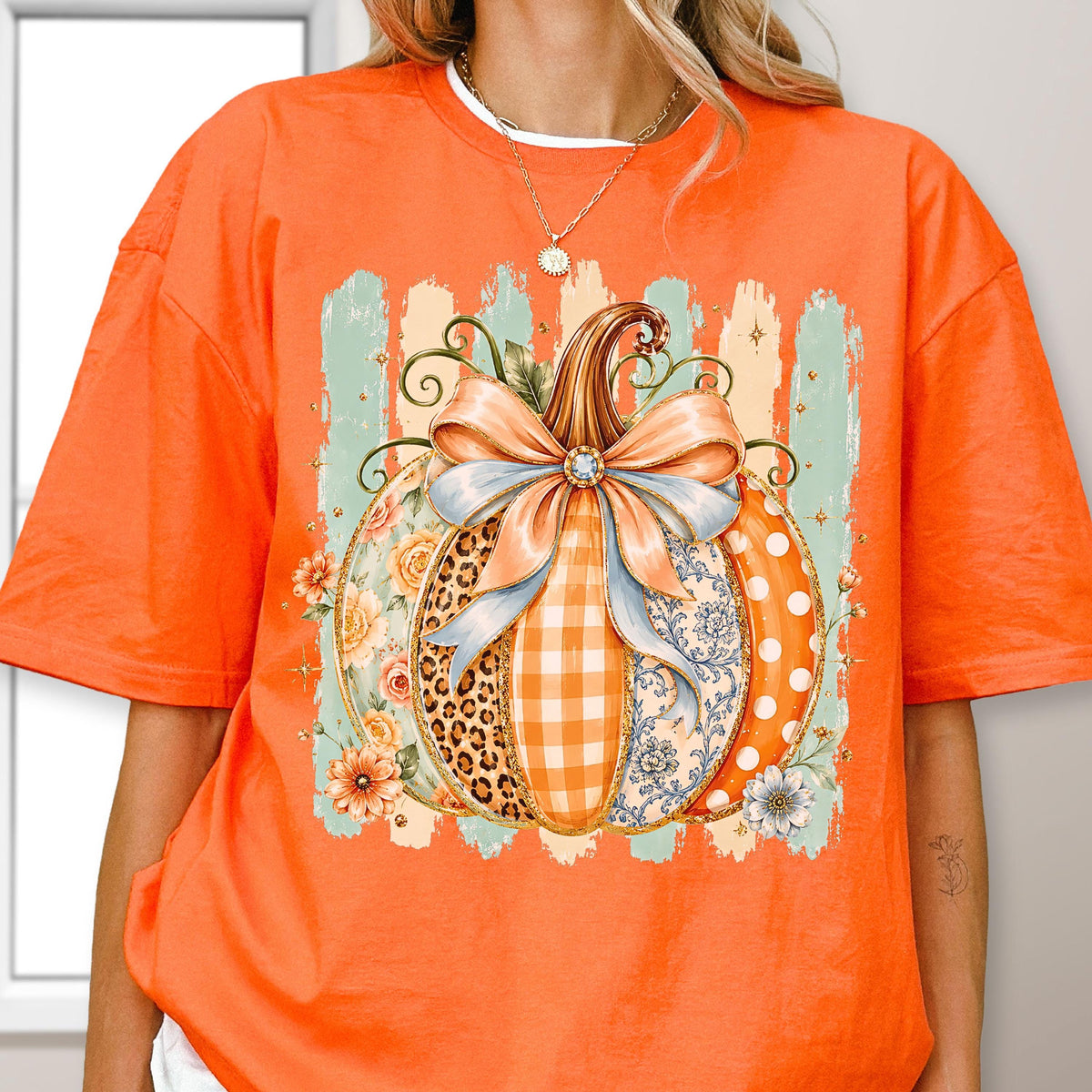 May include: An orange t-shirt featuring a large graphic of a pumpkin decorated with a light blue bow. The pumpkin design includes sections with leopard print, orange gingham, floral patterns, and white polka dots, set against a light green background.