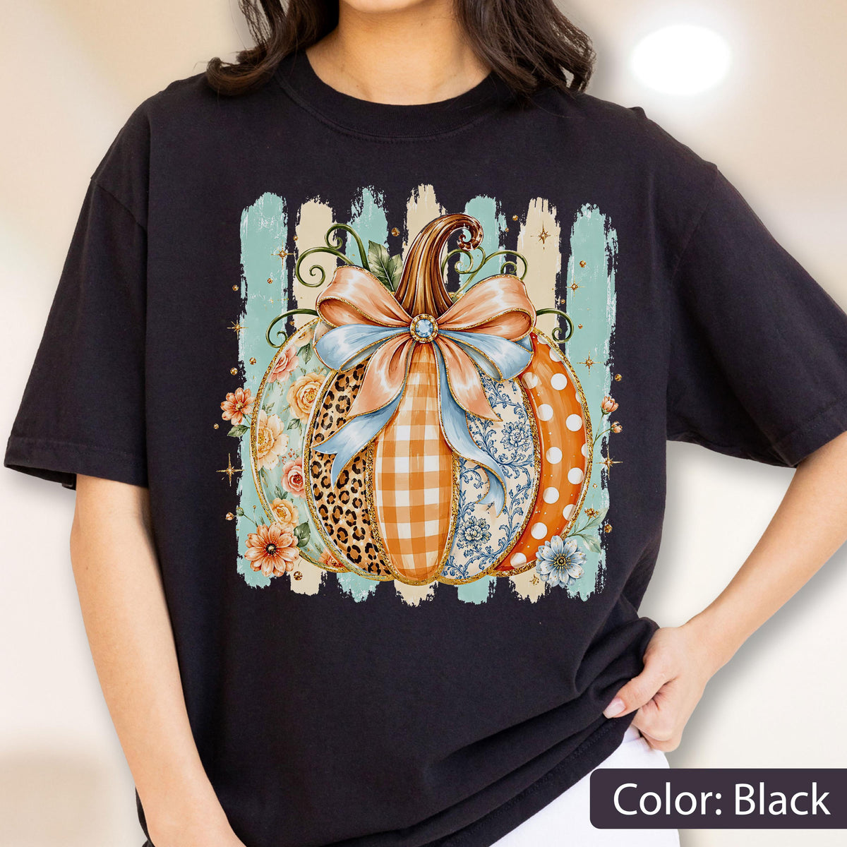May include: A black t-shirt featuring a central graphic of a pumpkin decorated with a large light blue bow. The pumpkin segments display various patterns including leopard print, orange gingham, floral motifs, and white polka dots on an orange background.