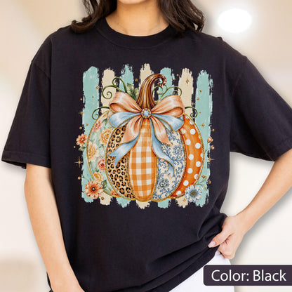 May include: A black t-shirt featuring a central graphic of a pumpkin decorated with a large light blue bow. The pumpkin segments display various patterns including leopard print, orange gingham, floral motifs, and white polka dots on an orange background.