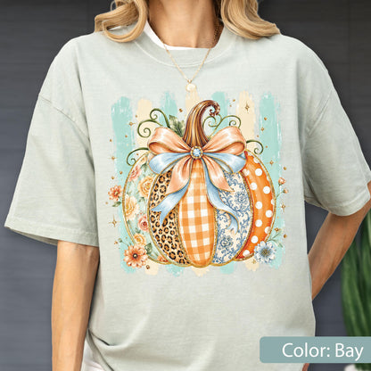 May include: A light sage green short-sleeve t-shirt featuring a central graphic of a pumpkin decorated with various patterns including leopard print, orange gingham, and floral motifs. A large light blue bow sits atop the pumpkin.