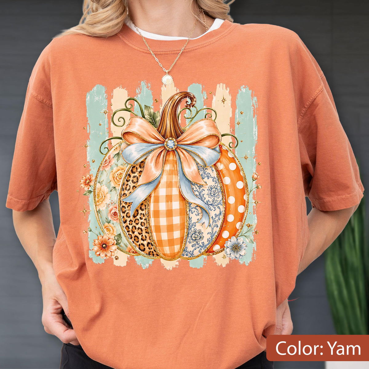 May include: A yam-colored t-shirt featuring a central graphic of a pumpkin decorated with a large light blue bow. The pumpkin segments display various patterns including leopard print, orange gingham, floral motifs, and white polka dots on orange.