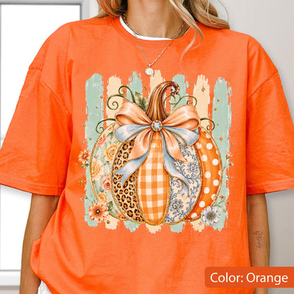 May include: An orange short-sleeve t-shirt featuring a large graphic of a pumpkin decorated with a light blue bow. The pumpkin design includes sections with leopard print, orange gingham, floral patterns, and white polka dots on an orange background.