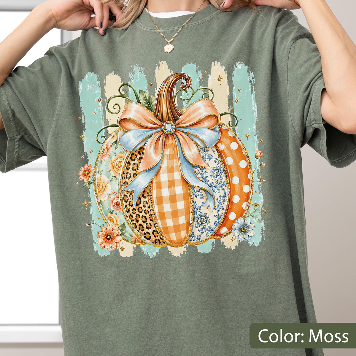 May include: A moss green t-shirt featuring a large central graphic of a pumpkin decorated with a light blue bow. The pumpkin segments display various patterns including leopard print, orange gingham, floral motifs, and white polka dots on orange.