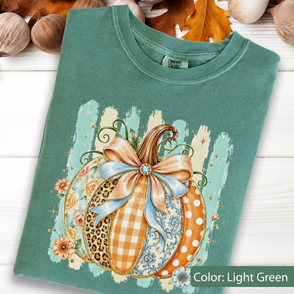 May include: A light green t-shirt featuring a graphic of a pumpkin decorated with various patterns including leopard print, orange gingham, blue floral, and white polka dots. The pumpkin is adorned with a large light blue bow and orange flowers.
