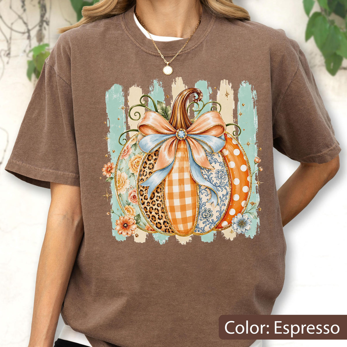 May include: An espresso-colored short-sleeve t-shirt featuring a central graphic of a pumpkin decorated with a large light blue bow. The pumpkin design incorporates various patterns including leopard print, orange gingham, floral motifs, and white polka dots.