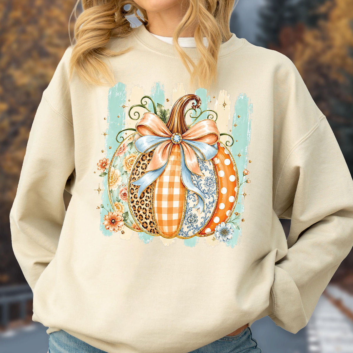 May include: A beige crewneck sweatshirt featuring a central graphic of a pumpkin decorated with various patterns including leopard print, orange gingham, blue floral, and white polka dots, topped with a large light blue bow and small flowers.
