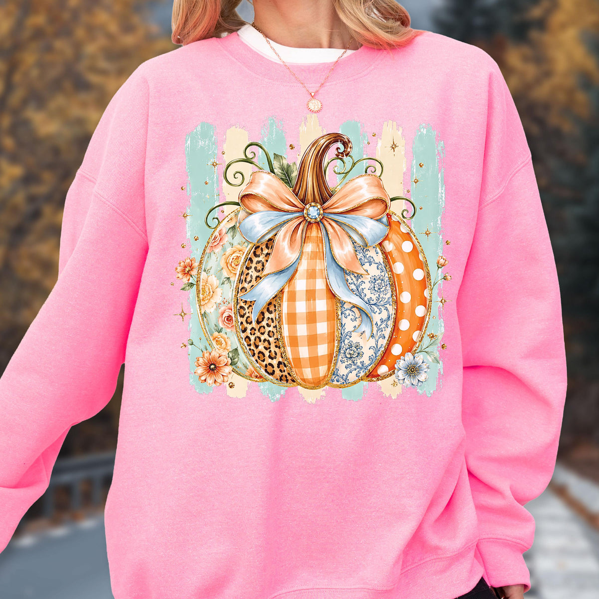 May include: A bright pink crewneck sweatshirt featuring a central graphic of a pumpkin decorated with various patterns including leopard print, orange gingham, and blue floral designs, topped with a large light blue bow and small flowers.