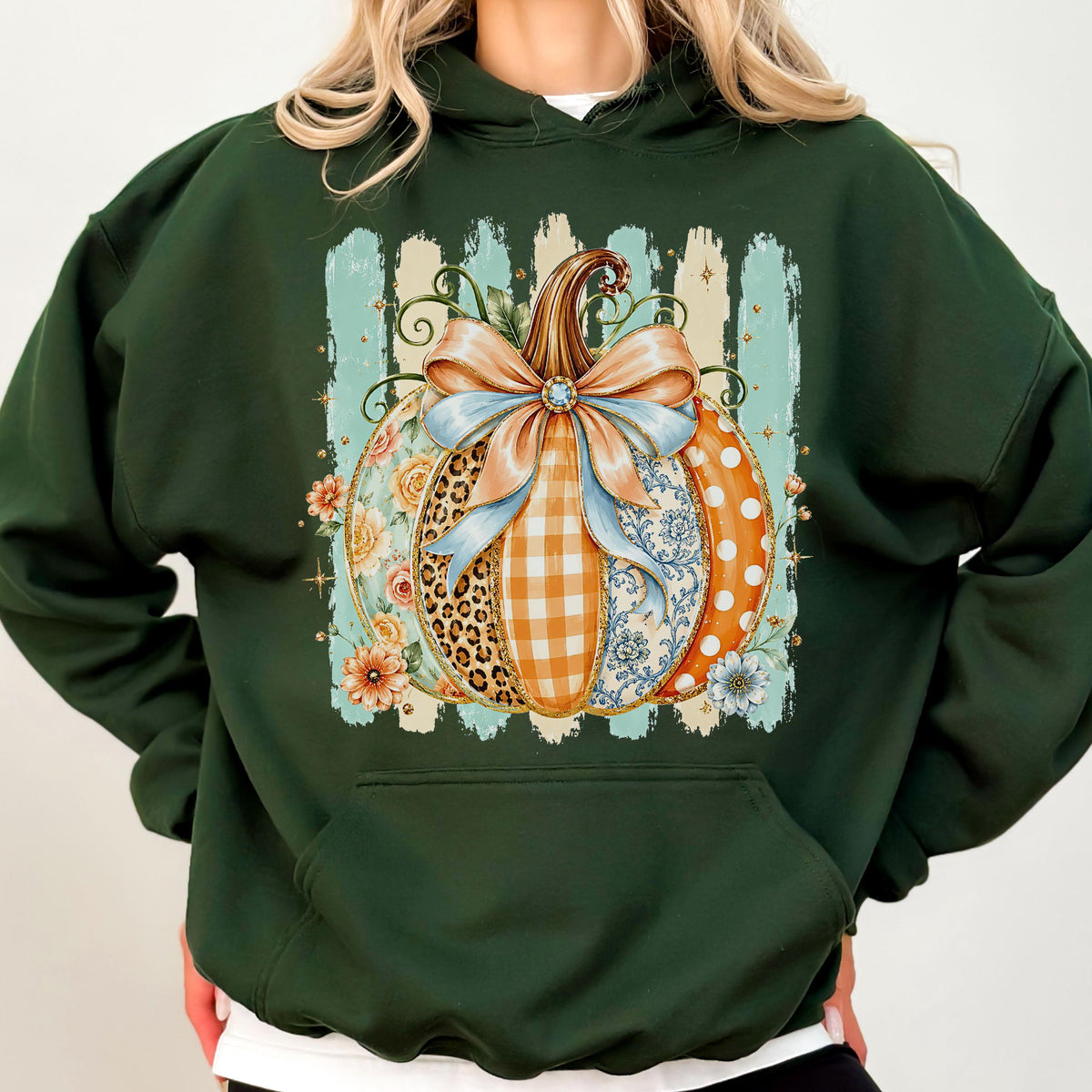 May include: A dark green hooded sweatshirt featuring a central graphic of a pumpkin decorated with various patterns including leopard print, orange gingham, and floral motifs, tied with a large light blue bow against a vertical striped background.