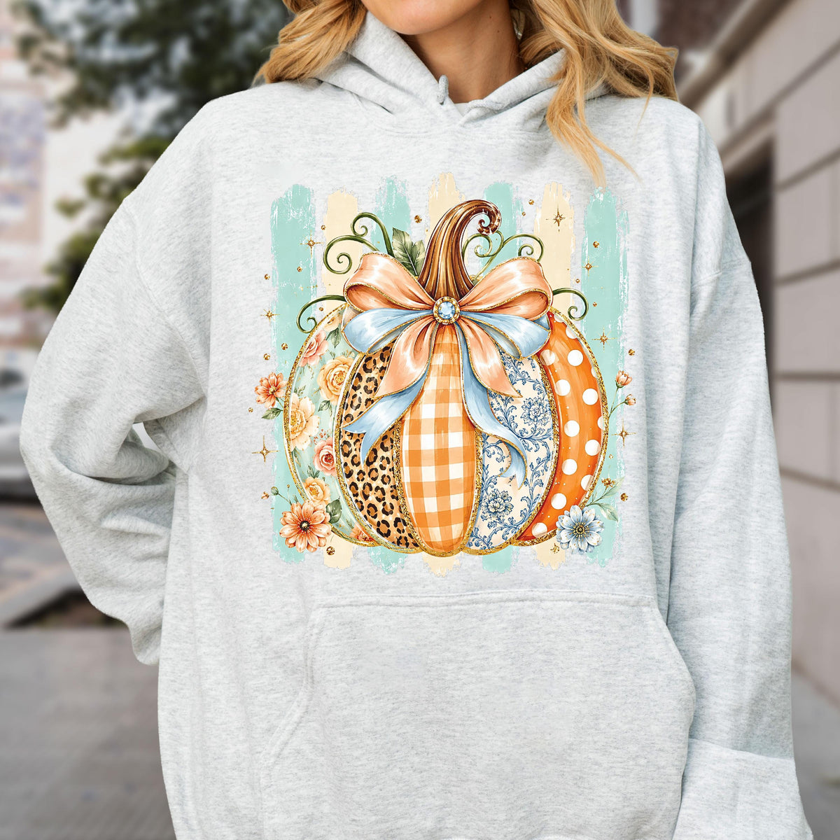 May include: A light gray hooded sweatshirt featuring a central graphic of a pumpkin decorated with various patterns including leopard print, orange gingham, and blue floral motifs. The pumpkin is topped with a large light blue bow and orange flowers.