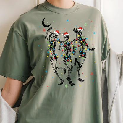 May include: An olive green t-shirt featuring a graphic print of three skeletons wearing red and white Santa hats. The skeletons are tangled in colorful Christmas lights, surrounded by small red, blue, and green snowflakes and a black crescent moon.