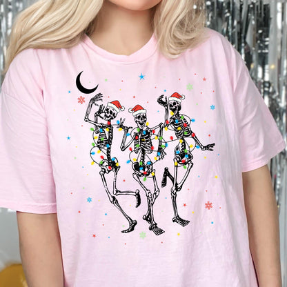May include: A light pink t-shirt featuring a graphic of three dancing skeletons wearing red and white Santa hats. The skeletons are wrapped in colorful Christmas lights, surrounded by small blue and red stars, snowflakes, and a black crescent moon.