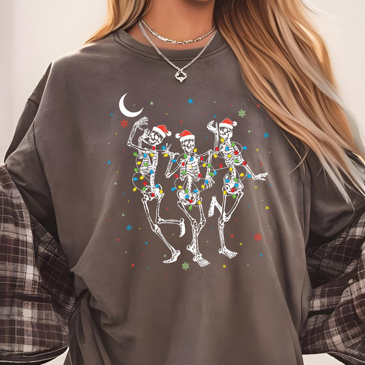 May include: A charcoal gray crewneck sweatshirt featuring a graphic of three white skeletons wearing red Santa hats and wrapped in colorful Christmas lights. The design includes a white crescent moon and small multi-colored star accents.