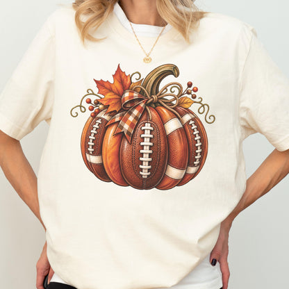 May include: A cream-colored t-shirt featuring a graphic of a pumpkin designed with the texture and white laces of a brown football. The pumpkin is decorated with a plaid ribbon bow, an orange maple leaf, and small red berries on its stem.