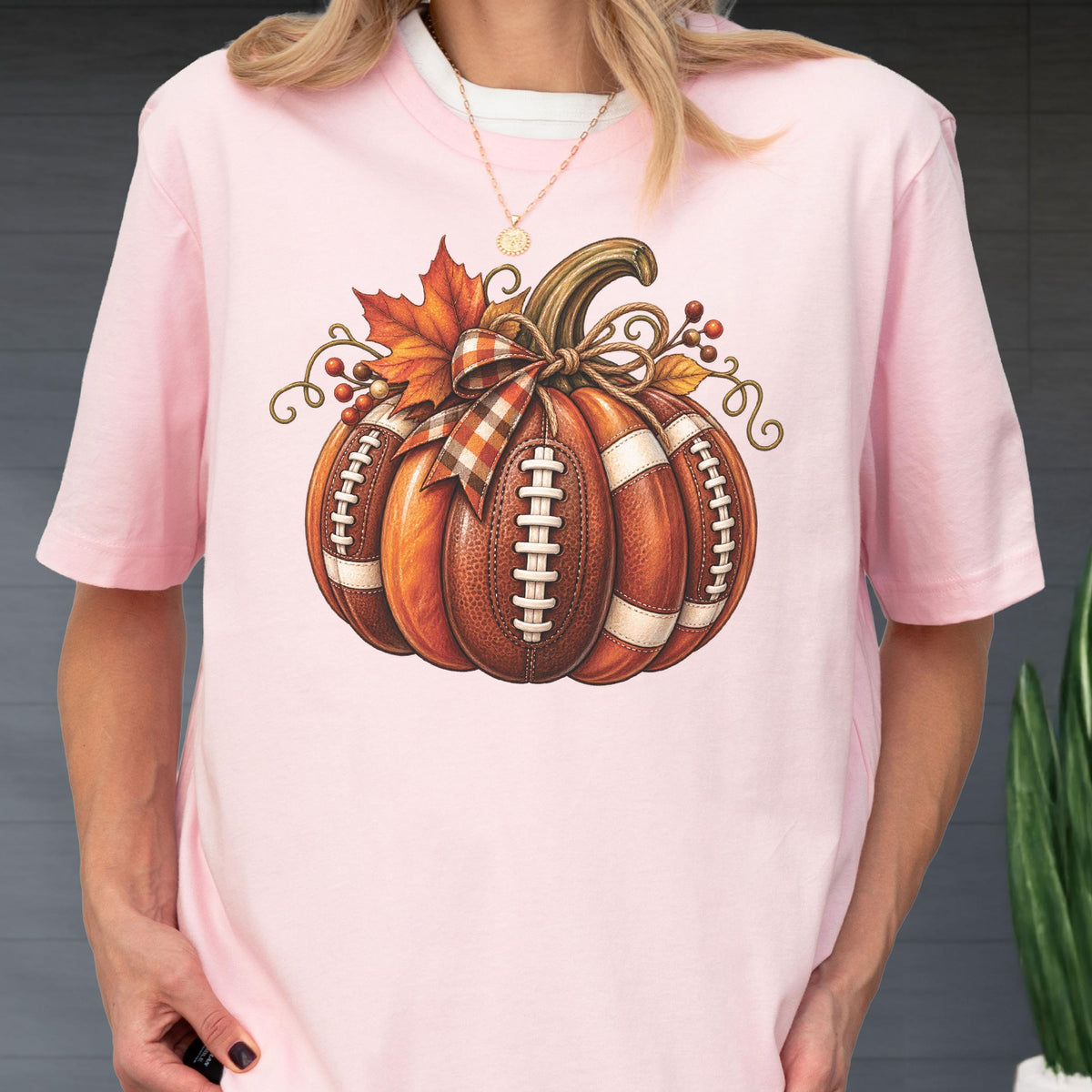 May include: A light pink short-sleeve t-shirt featuring a graphic of a pumpkin designed with the texture and white laces of an American football. The pumpkin is decorated with a plaid bow, an orange maple leaf, and small brown berries.