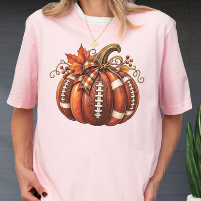 May include: A light pink short-sleeve t-shirt featuring a graphic of a pumpkin designed with the texture and white laces of an American football. The pumpkin is decorated with a plaid bow, an orange maple leaf, and small brown berries.