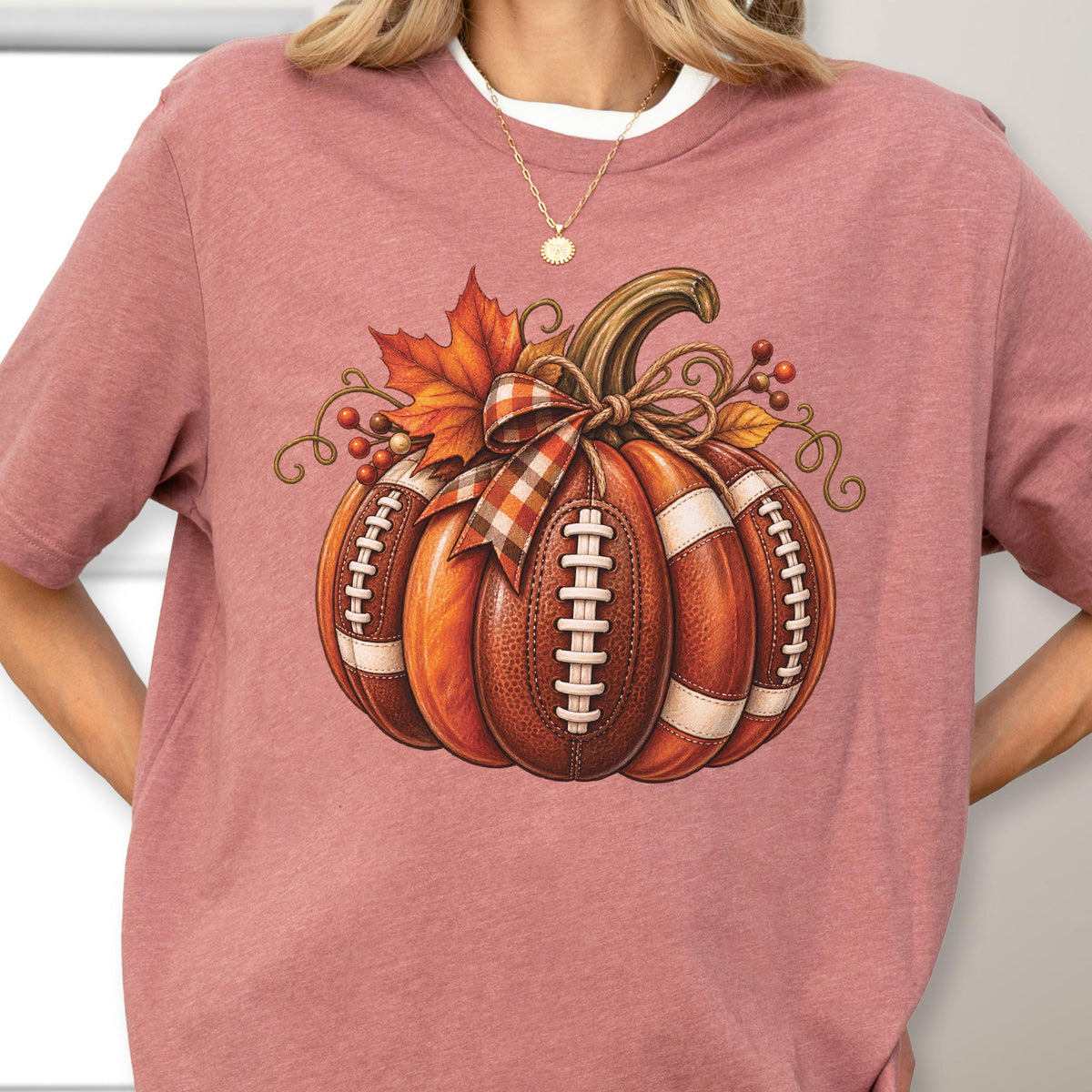 May include: A dusty rose short-sleeve t-shirt featuring a graphic of a pumpkin designed with the texture and white laces of an American football. The pumpkin is decorated with a plaid ribbon, an orange maple leaf, and small red berries.