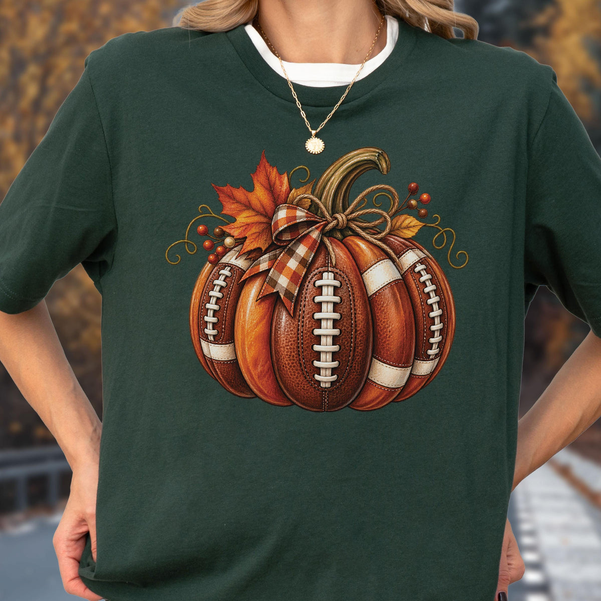 May include: A dark green t-shirt featuring a graphic of a pumpkin constructed from brown footballs with white laces. The pumpkin is decorated with a plaid ribbon bow, orange maple leaves, and small red berries, creating a seasonal autumn design.