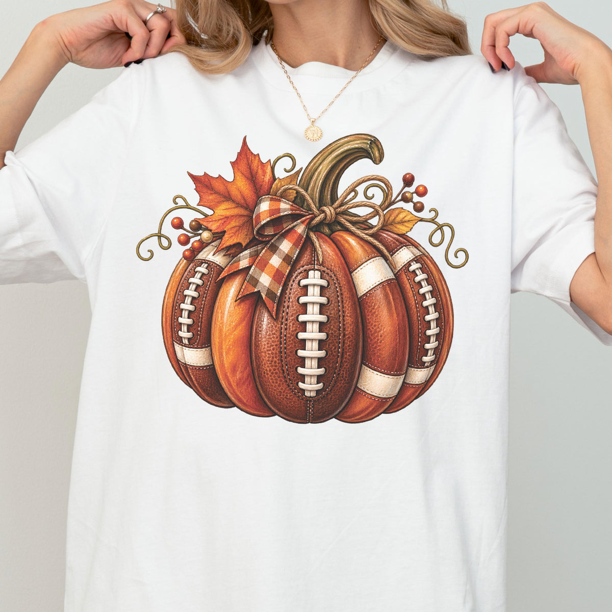 May include: A white t-shirt featuring a graphic of a pumpkin designed with the texture and white laces of an American football. The pumpkin is decorated with a plaid ribbon bow, an orange maple leaf, and small red berries on curly brown vines.