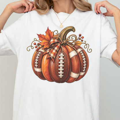 May include: A white t-shirt featuring a graphic of a pumpkin designed with the texture and white laces of an American football. The pumpkin is decorated with a plaid ribbon bow, an orange maple leaf, and small red berries on curly brown vines.