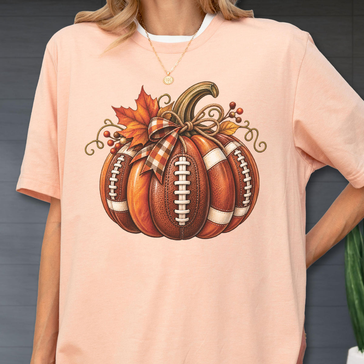May include: A peach-colored t-shirt featuring a graphic of a pumpkin designed with the texture and white laces of an American football. The pumpkin is decorated with a plaid ribbon bow, a single orange maple leaf, and small red berries.