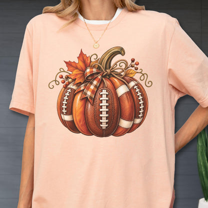 May include: A peach-colored t-shirt featuring a graphic of a pumpkin designed with the texture and white laces of an American football. The pumpkin is decorated with a plaid ribbon bow, a single orange maple leaf, and small red berries.