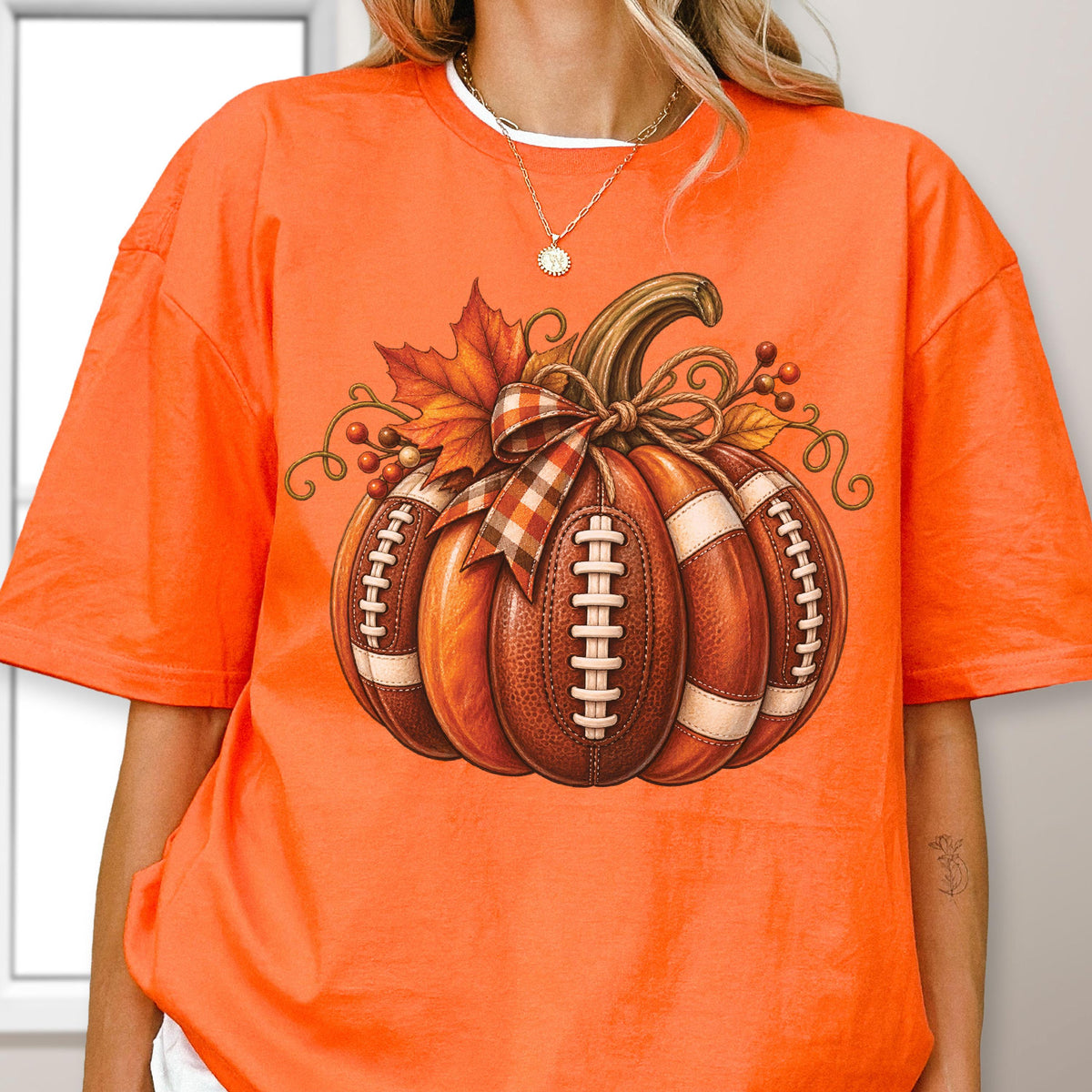 May include: A bright orange short-sleeve t-shirt featuring a large graphic of a pumpkin designed with the texture and white laces of an American football. The pumpkin is decorated with a plaid ribbon, autumn leaves, and small brown berries.
