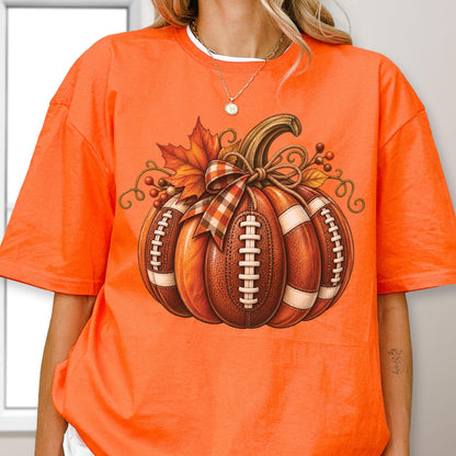 May include: A bright orange short-sleeve t-shirt featuring a large graphic of a pumpkin designed with the texture and white laces of an American football. The pumpkin is decorated with a plaid ribbon, autumn leaves, and small brown berries.