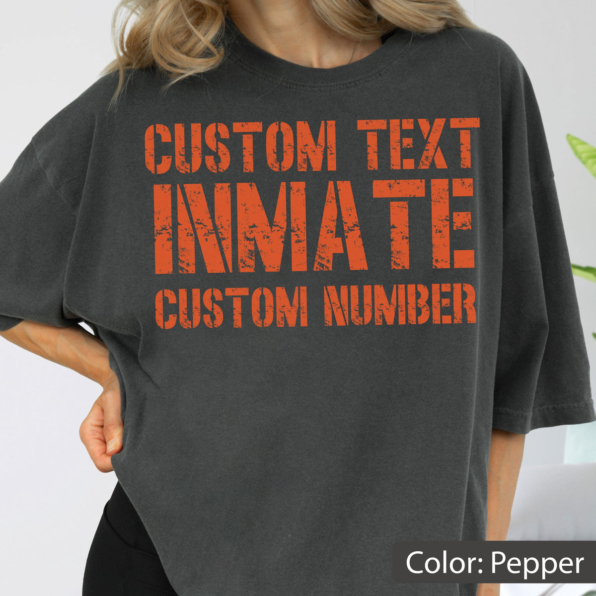 May include: A pepper-colored short-sleeve t-shirt featuring distressed orange block lettering on the front. The text reads CUSTOM TEXT, INMATE, and CUSTOM NUMBER in a bold, weathered font style across the chest area of the garment.