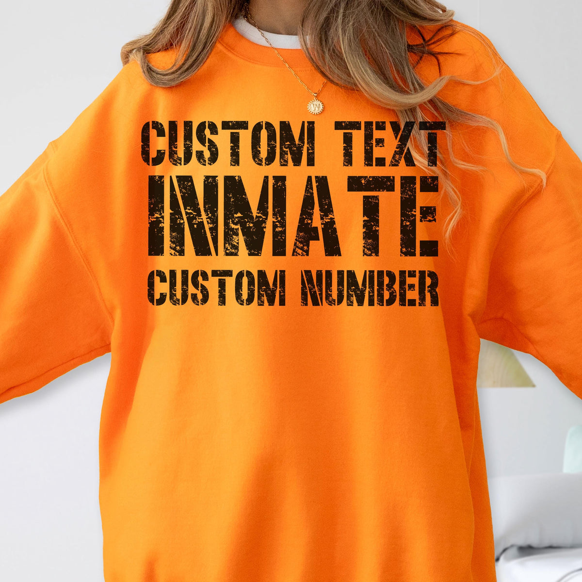 May include: A bright orange crewneck sweatshirt featuring black distressed-style text printed on the chest. The text reads CUSTOM TEXT, INMATE, and CUSTOM NUMBER in a bold, blocky font. The garment has long sleeves and a ribbed neckline.