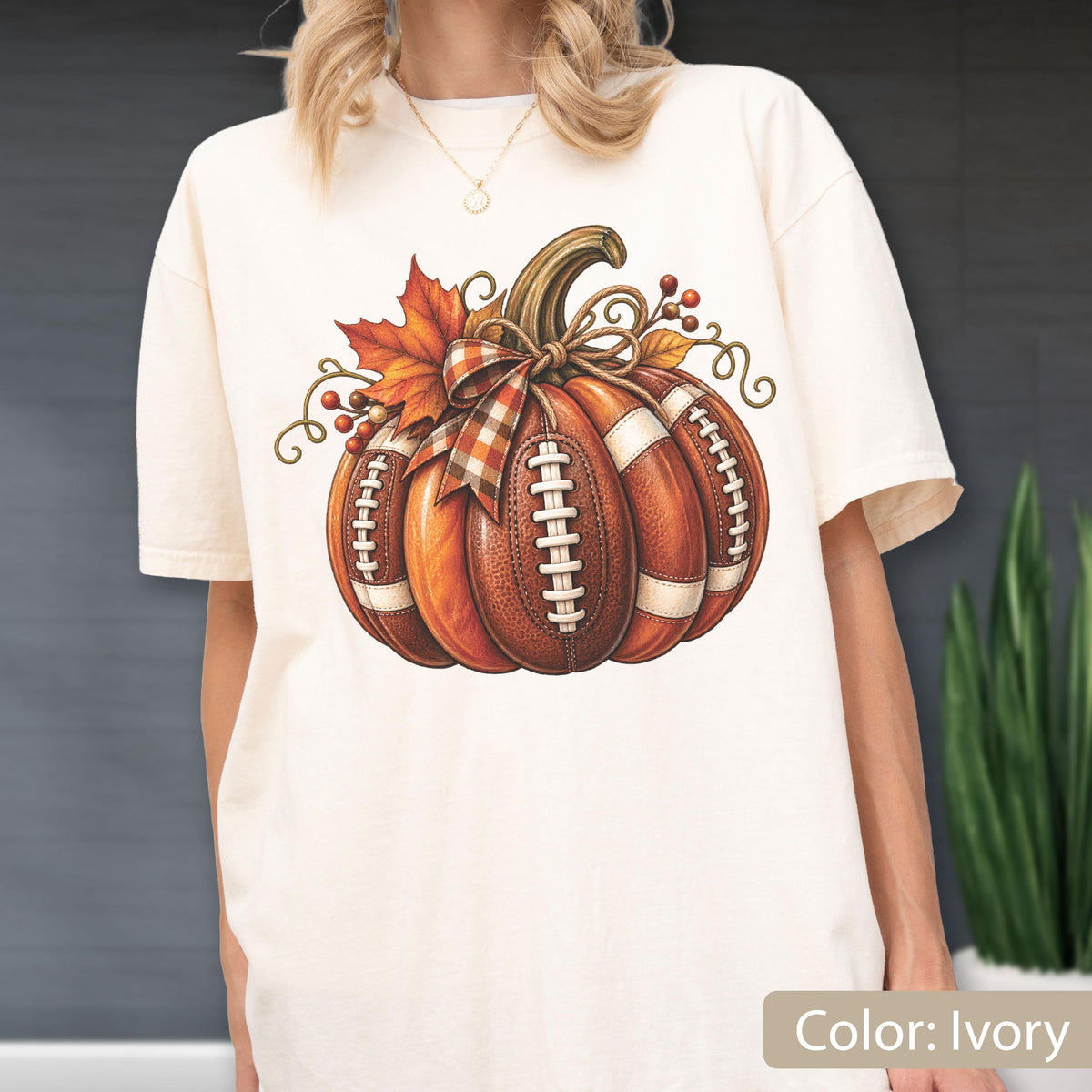 May include: An ivory short-sleeve t-shirt featuring a graphic of a pumpkin designed to look like a brown leather football with white laces. The pumpkin is decorated with a plaid bow, orange maple leaves, and small red berries on its stem.