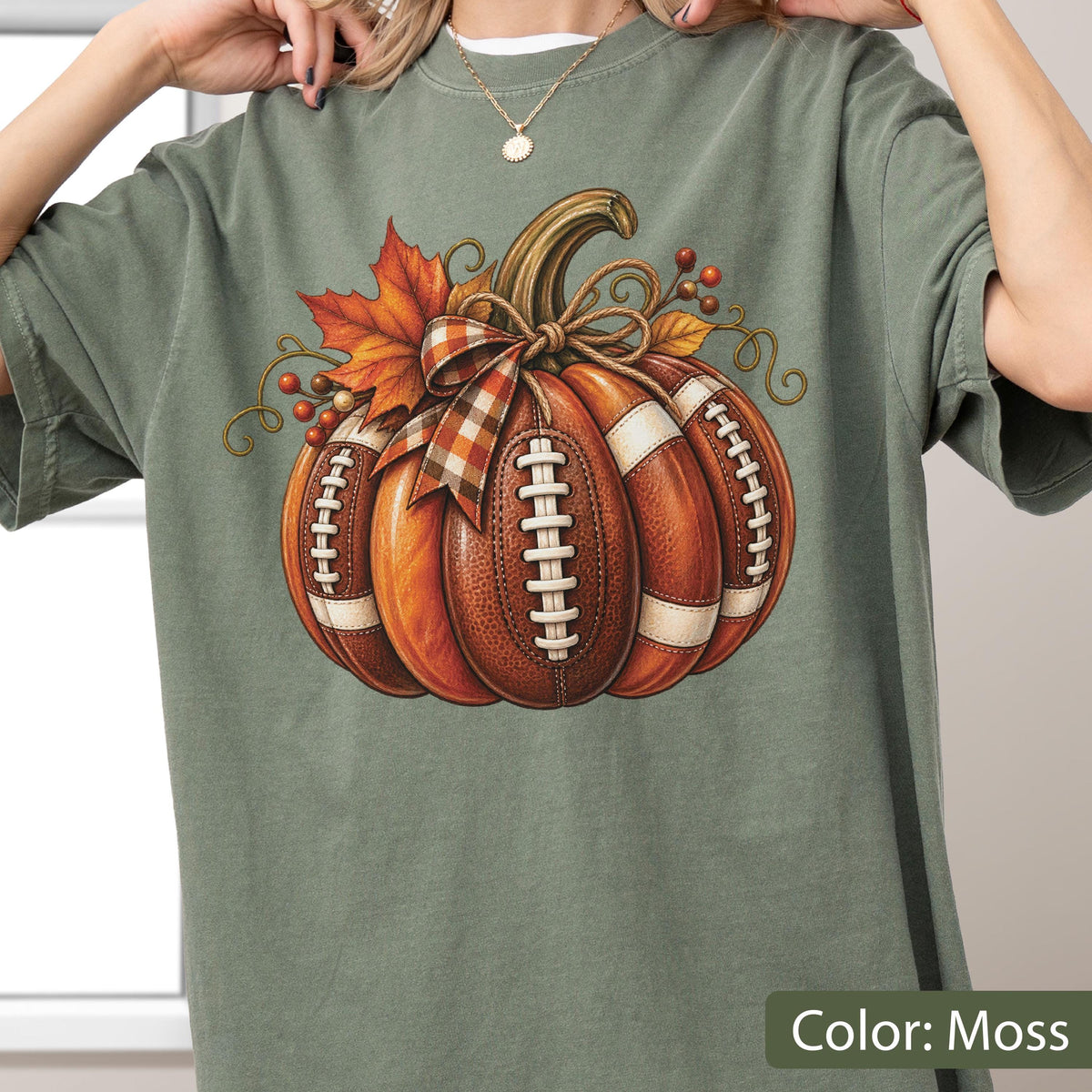 May include: A moss green t-shirt featuring a graphic of a pumpkin designed with the texture and white laces of a football. The pumpkin is decorated with a plaid ribbon, orange maple leaves, and small brown berries, creating a seasonal autumn theme.