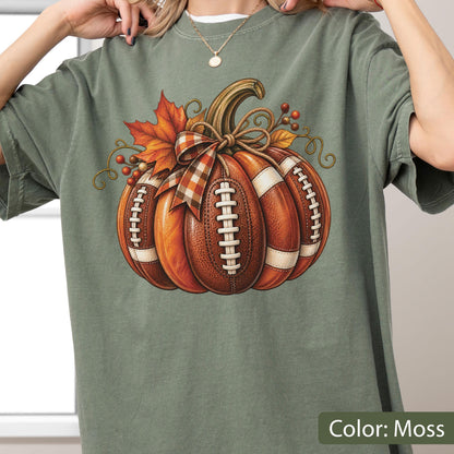 May include: A moss green t-shirt featuring a graphic of a pumpkin designed with the texture and white laces of a football. The pumpkin is decorated with a plaid ribbon, orange maple leaves, and small brown berries, creating a seasonal autumn theme.