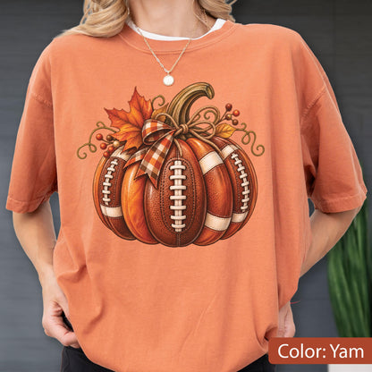 May include: A yam-colored short-sleeve t-shirt featuring a central graphic of a pumpkin designed with the texture and white laces of an American football. The pumpkin is decorated with a plaid ribbon bow, orange maple leaves, and small berries.