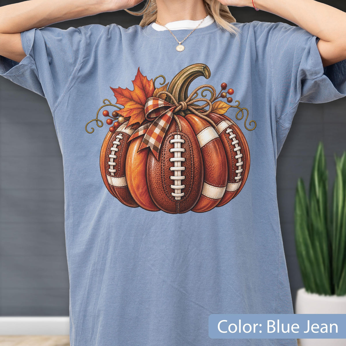 May include: A blue jean colored t-shirt featuring a graphic of a pumpkin designed with the texture and white laces of a football. The pumpkin is decorated with a plaid ribbon bow, an orange maple leaf, and small red berries on a vine.