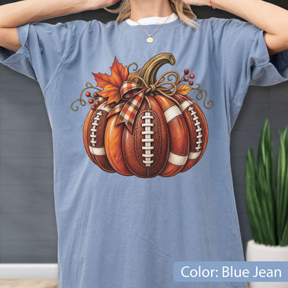 May include: A blue jean colored t-shirt featuring a graphic of a pumpkin designed with the texture and white laces of a football. The pumpkin is decorated with a plaid ribbon bow, an orange maple leaf, and small red berries on a vine.