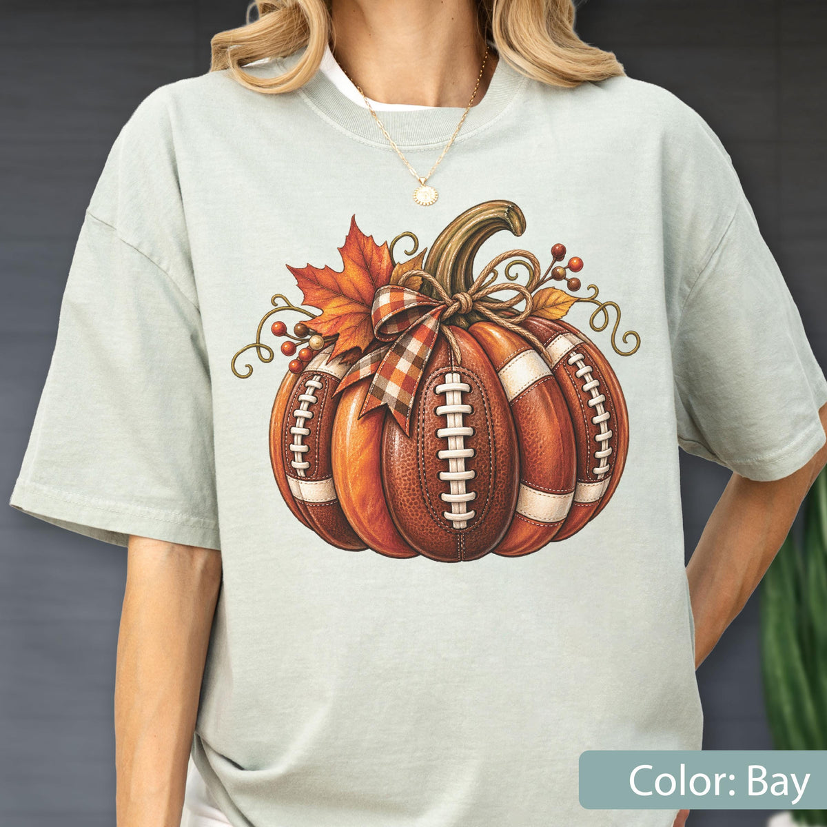 May include: A light sage green t-shirt featuring a central graphic of a pumpkin designed with the texture and white laces of an American football. The pumpkin is decorated with a plaid ribbon, autumn leaves, and small red berries.