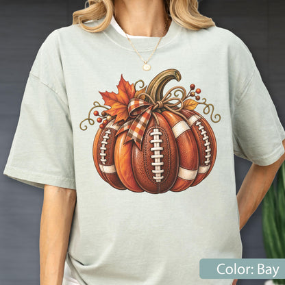 May include: A light sage green t-shirt featuring a central graphic of a pumpkin designed with the texture and white laces of an American football. The pumpkin is decorated with a plaid ribbon, autumn leaves, and small red berries.