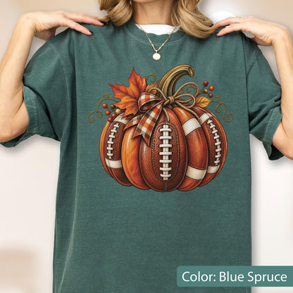 May include: A blue spruce t-shirt featuring a central graphic of a pumpkin designed with the texture and white laces of an American football. The pumpkin is decorated with a plaid ribbon bow, orange autumn leaves, and small red berries.