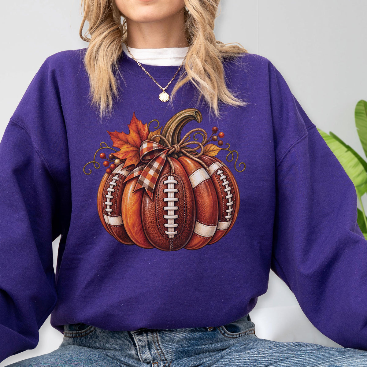 May include: A deep purple long-sleeve sweatshirt featuring a central graphic of a pumpkin designed with the texture and white laces of an American football. The pumpkin is accented with a plaid ribbon bow, orange maple leaves, and small red berries.
