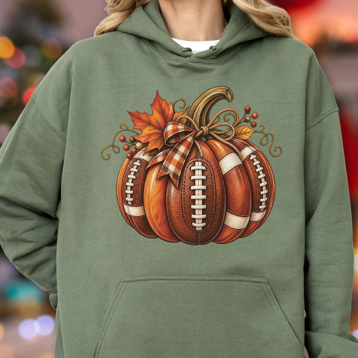 May include: An olive green hooded sweatshirt featuring a central graphic of a pumpkin designed with the texture and white laces of an American football. The pumpkin is decorated with a plaid ribbon bow, orange maple leaves, and red berries.