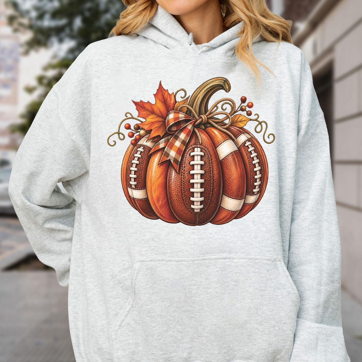 May include: A light gray hooded sweatshirt featuring a central graphic of a pumpkin designed with the texture and white laces of an American football. The pumpkin is decorated with a plaid ribbon bow, an orange maple leaf, and brown berries.