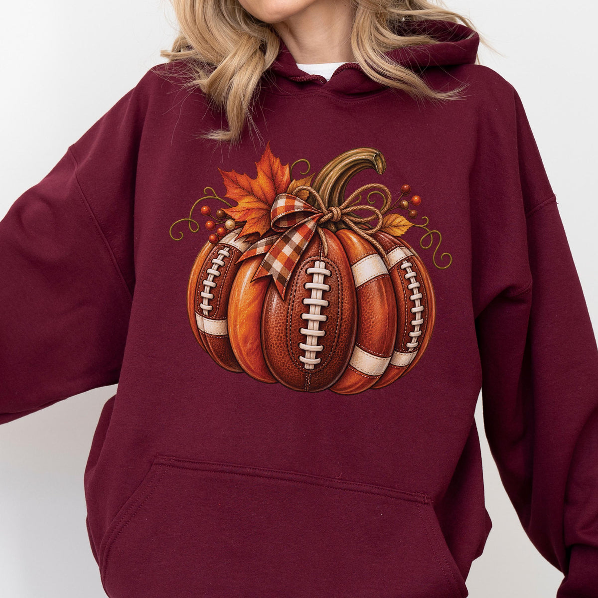 May include: A maroon hooded sweatshirt featuring a graphic of a pumpkin designed to look like a brown leather football with white laces. The pumpkin is decorated with a plaid ribbon bow, orange maple leaves, and small red berries on the top.