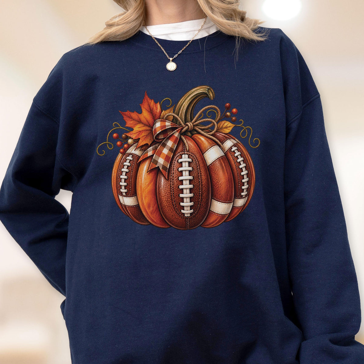 May include: A navy blue crewneck sweatshirt featuring a central graphic of a pumpkin designed with the texture and white laces of an American football. The pumpkin is adorned with a plaid ribbon bow, orange maple leaves, and small red berries.