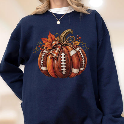 May include: A navy blue crewneck sweatshirt featuring a central graphic of a pumpkin designed with the texture and white laces of an American football. The pumpkin is adorned with a plaid ribbon bow, orange maple leaves, and small red berries.