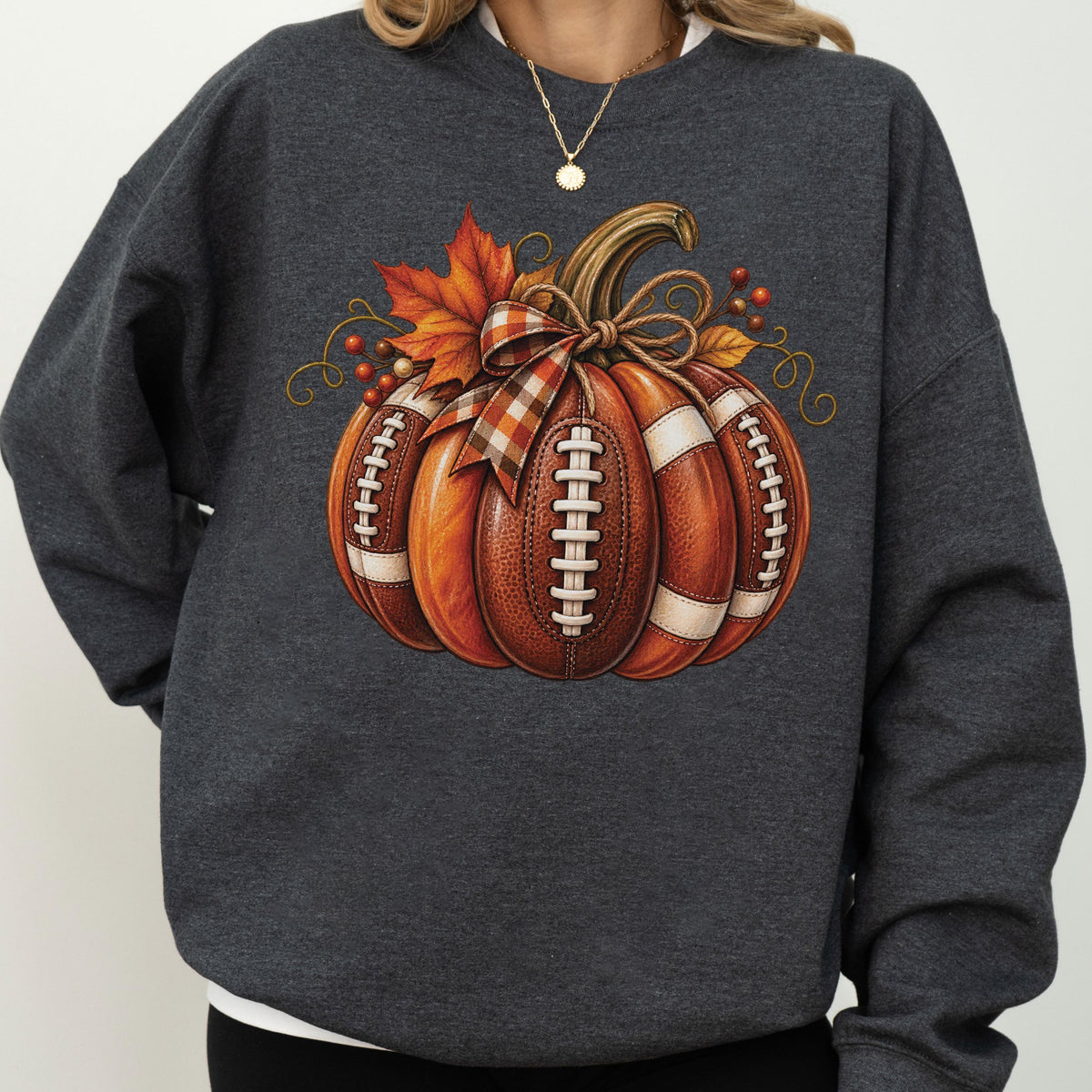 May include: A dark charcoal gray crewneck sweatshirt featuring a central graphic of a pumpkin designed with the texture and white laces of an American football. The pumpkin is adorned with a plaid ribbon bow, orange maple leaves, and small red berries.