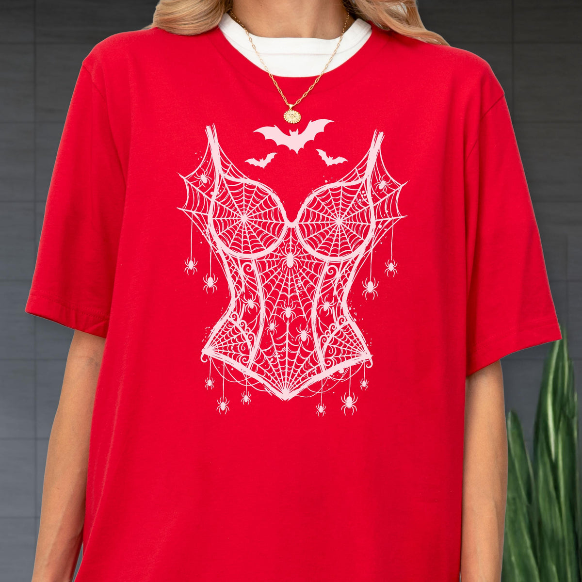May include: A bright red short-sleeved t-shirt featuring a white graphic print of a corset made from spiderwebs. The design includes small spiders hanging from the webbed corset and three bats flying above the neckline. A gold chain necklace is worn.