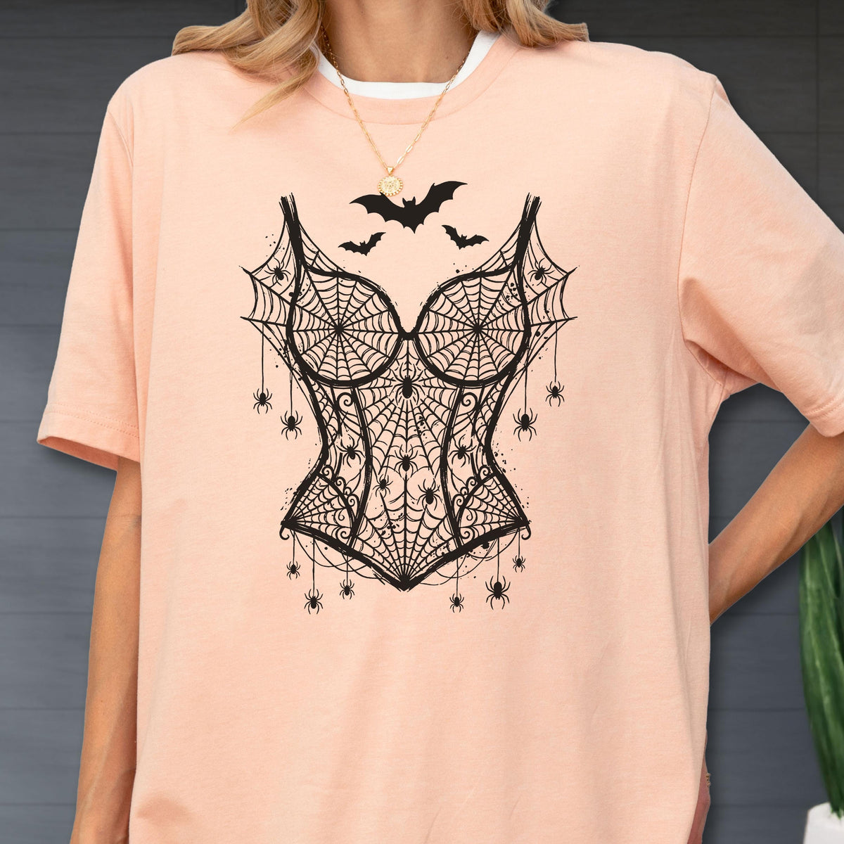 May include: A peach-colored short-sleeve t-shirt featuring a black graphic print of a corset made of spiderwebs and spiders, with three small bats flying above the corset design. The shirt has a crew neckline and a relaxed, loose-fitting cut.