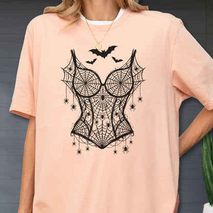 May include: A peach-colored short-sleeve t-shirt featuring a black graphic print of a corset made of spiderwebs and spiders, with three small bats flying above the corset design. The shirt has a crew neckline and a relaxed, loose-fitting cut.
