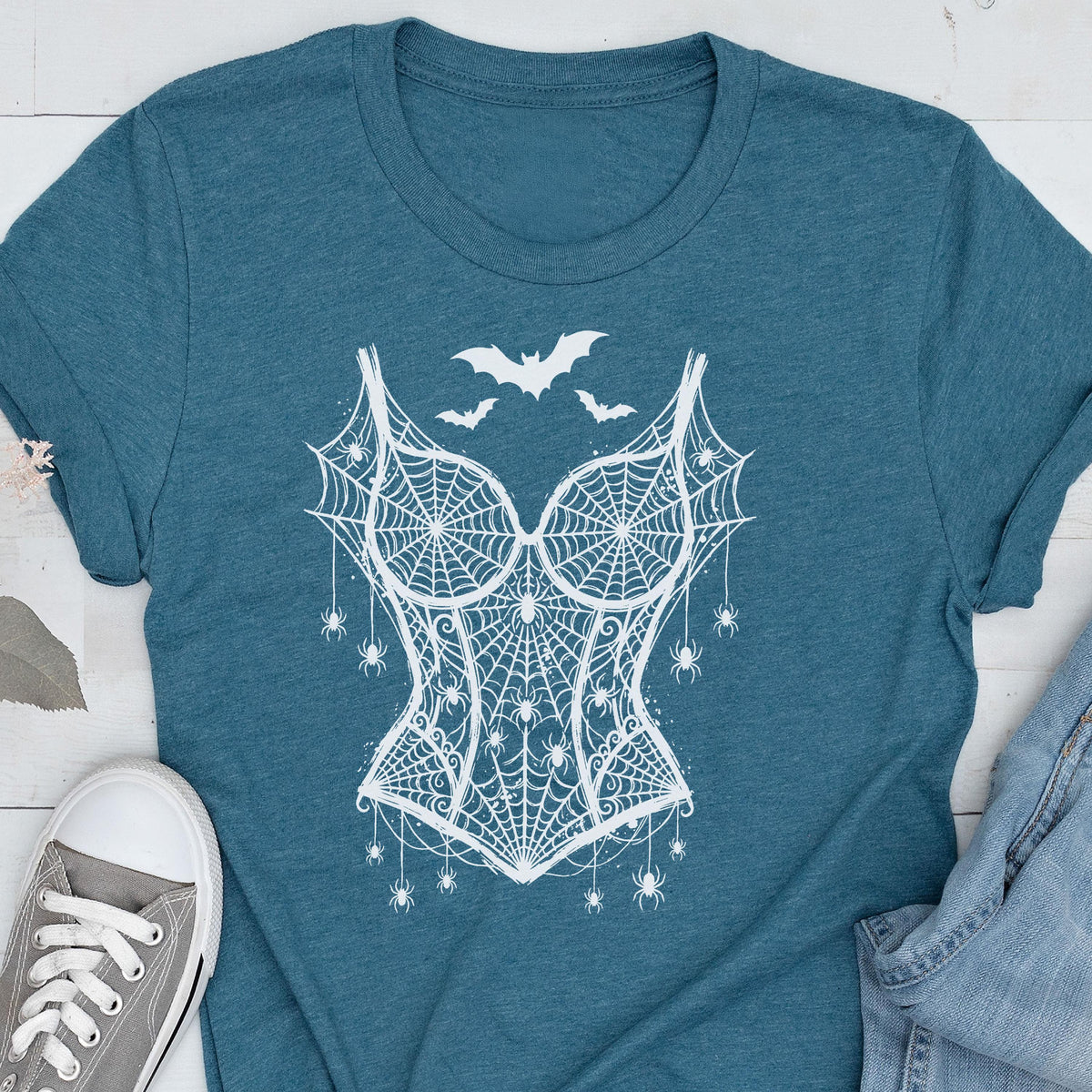 May include: A teal short-sleeved t-shirt featuring a white graphic print of a corset made from spiderwebs, adorned with small hanging spiders and three bats flying above the bodice design, centered on the front of the garment.