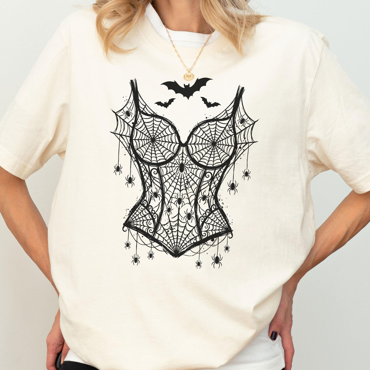 May include: A cream-colored short-sleeve t-shirt featuring a black graphic print of a corset made of spiderwebs. The design includes multiple small spiders hanging from the webbed corset and three black bats flying above the neckline.